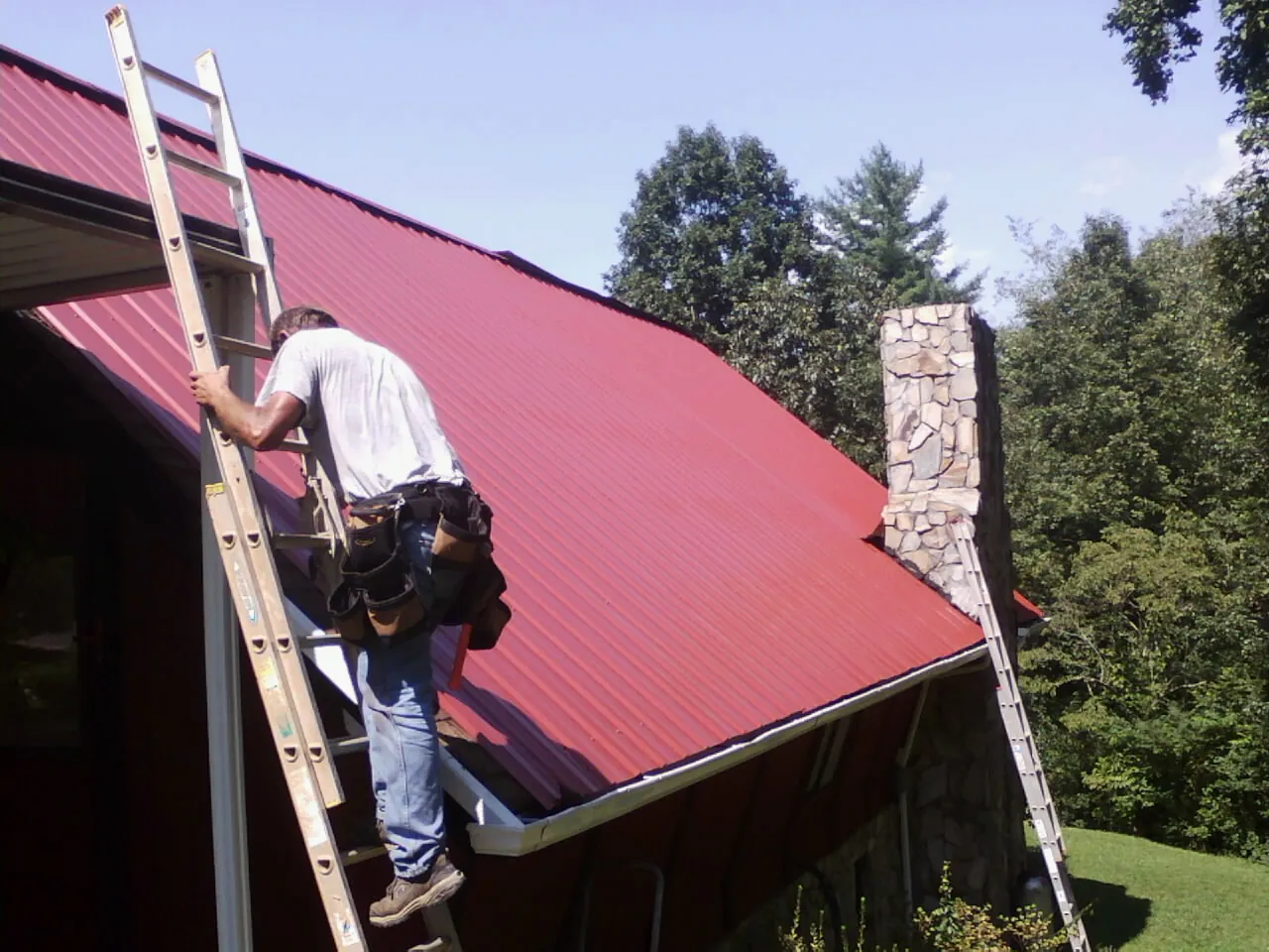 Roofer on a ladder beside a red metal roof and stone chimney — Soffit & Fascia Repair by Roofing Brothers in Posen